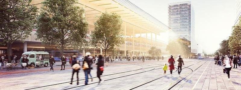 Ett stort, öppet torg där människor promenerar. I bakgrunden syns stationen.