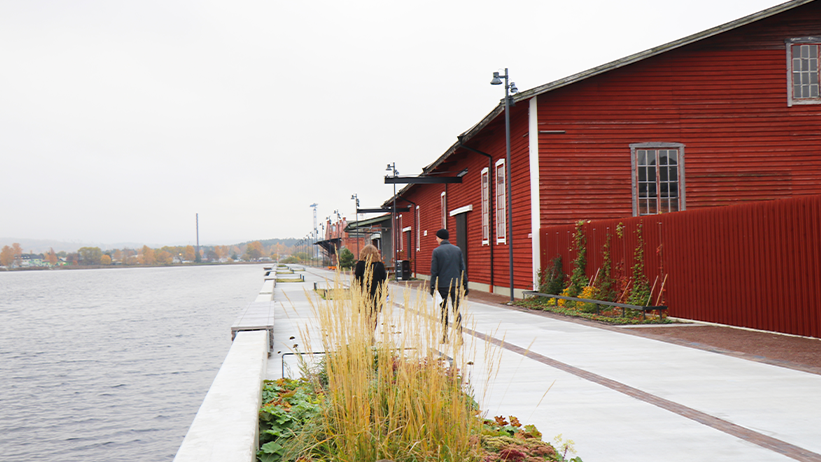 Ett rött trähus till höger i bild. Till vänster i bild en kaj i ljus sten som går längs vattnet. Två personer går på kajen.