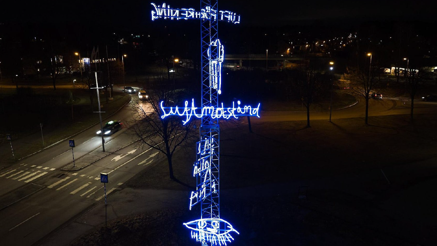 En ljuskonstverk som sitter monterat på en mastliknande ställning. Verket består av blåa lysande neonslingor. Texten "luftmotstånd" syns.