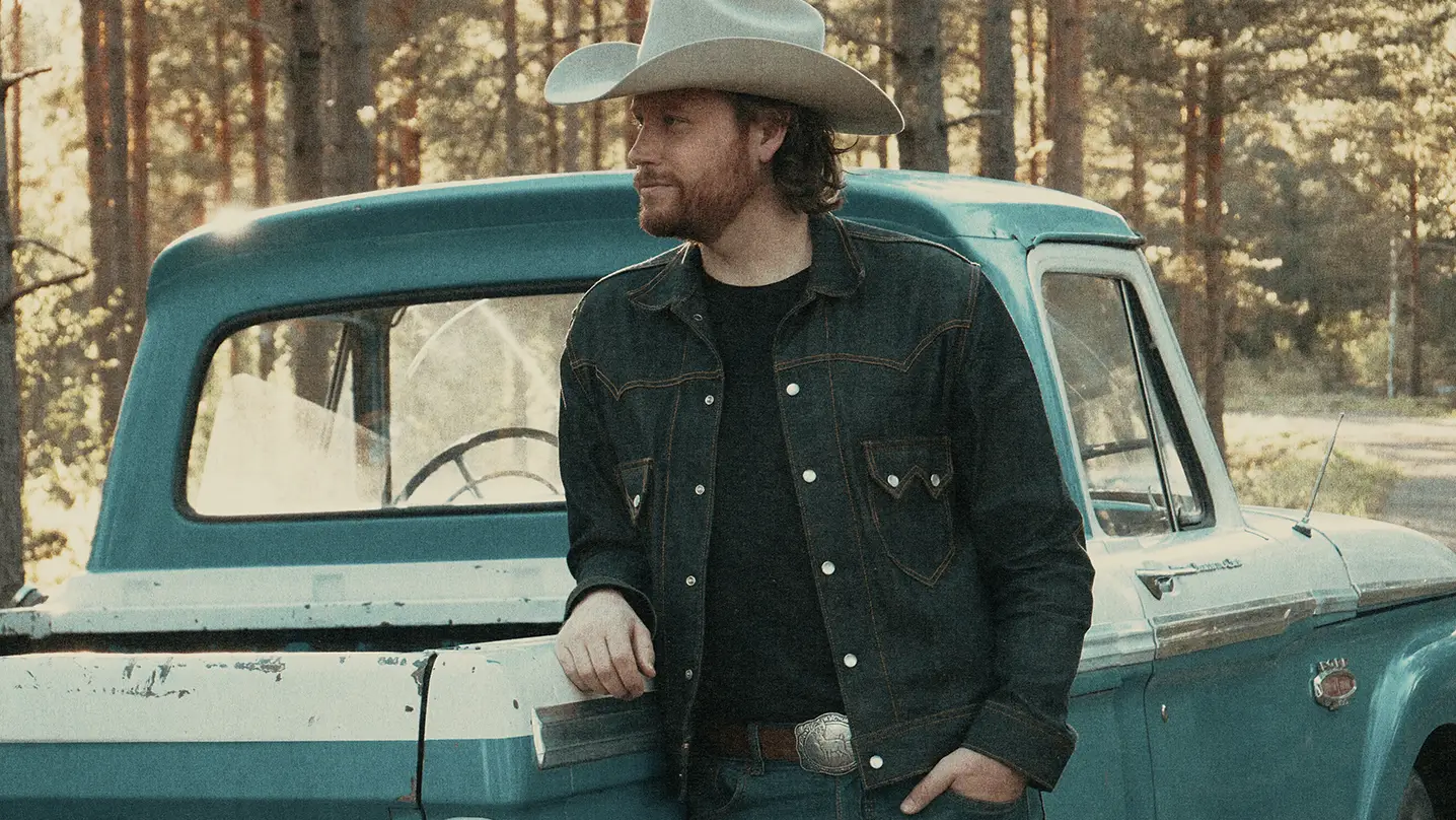 En man med jeansjacka och vit cowboyhatt står lutad mot en turkos pickup.