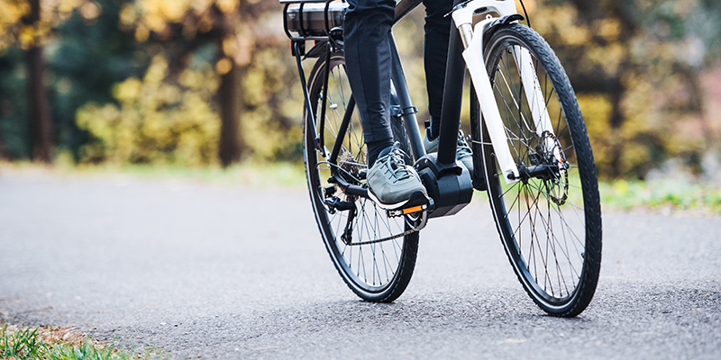 Närbild på nedre delen av en cykel som åker på asfaltsväg. I bakgrunden syns en del växtlighet.