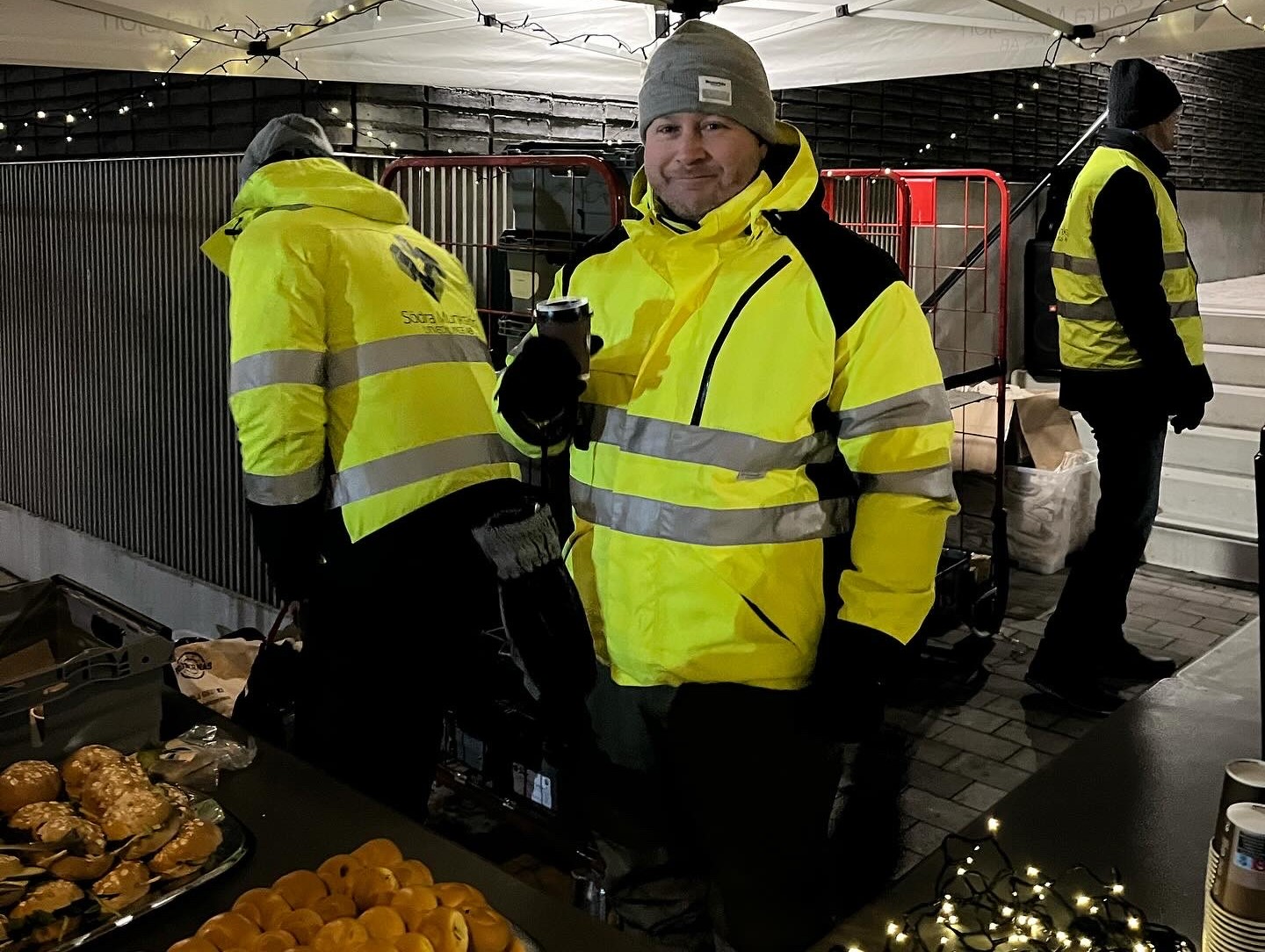 Emil som arbetar på SMUAB står i ett tält med en kopp kaffe i handen. Han ler och är klädd i gul varseljacka.