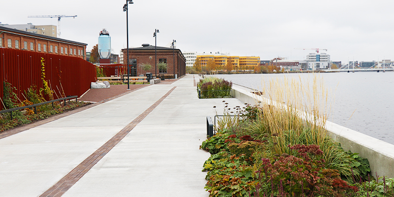 Falurött trästaket, stenbelagd kaj med tegeldetaljer, vackra höstplanteringar och gamla tegelbyggnader. I bakgrunden skymtar högskolan i Jönköping. 