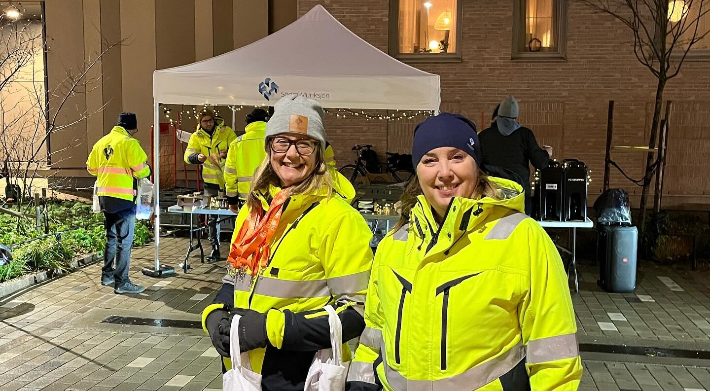 Anna och Jenny som arbetar på SMUAB står framför kameran och ler. De har på sig varseljackor och mössa och håller i de påsar som delades ut under inflyttningsfesten. I bakgrunden står ett vitt tält med SMUAB:s logga. Tältet är klätt med ljusslingor och i tältet står bor uppdukade med frukost.