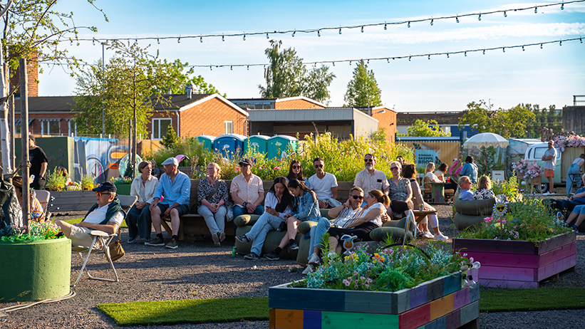 Människor sitter på bänkar bland grönska och ljusslingor i sommarsolen.