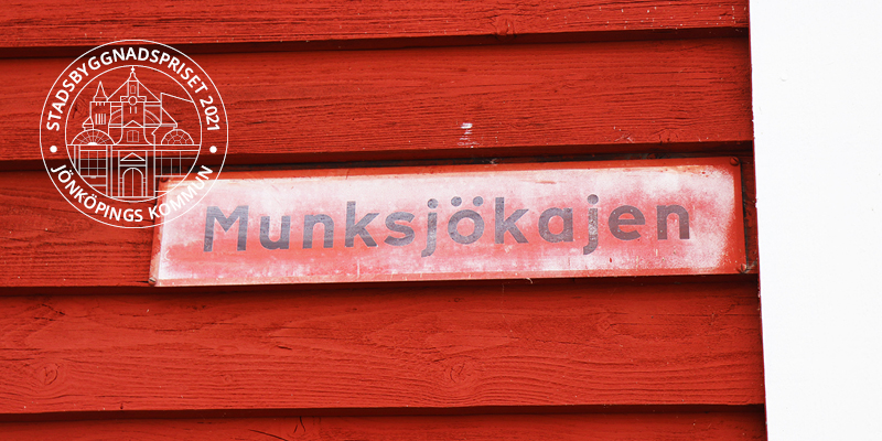 En gammal gatuskylt med text "Munksjökajen" på en faluröd trävägg.