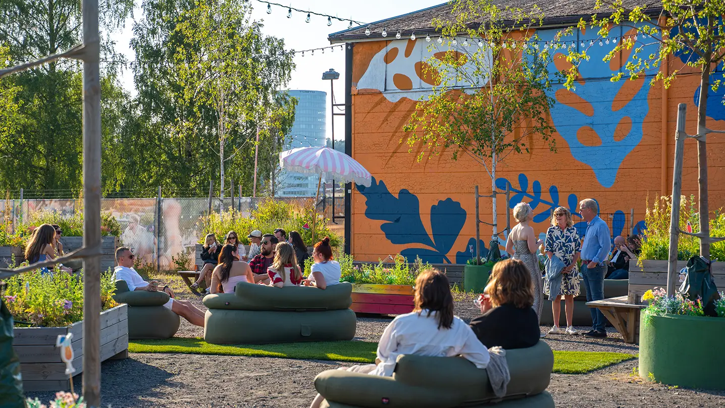 Människor sitter i gröna soffor på en prunkande grön innergård. Solen lyser och i luften hänger ljusslingor som ett tak.