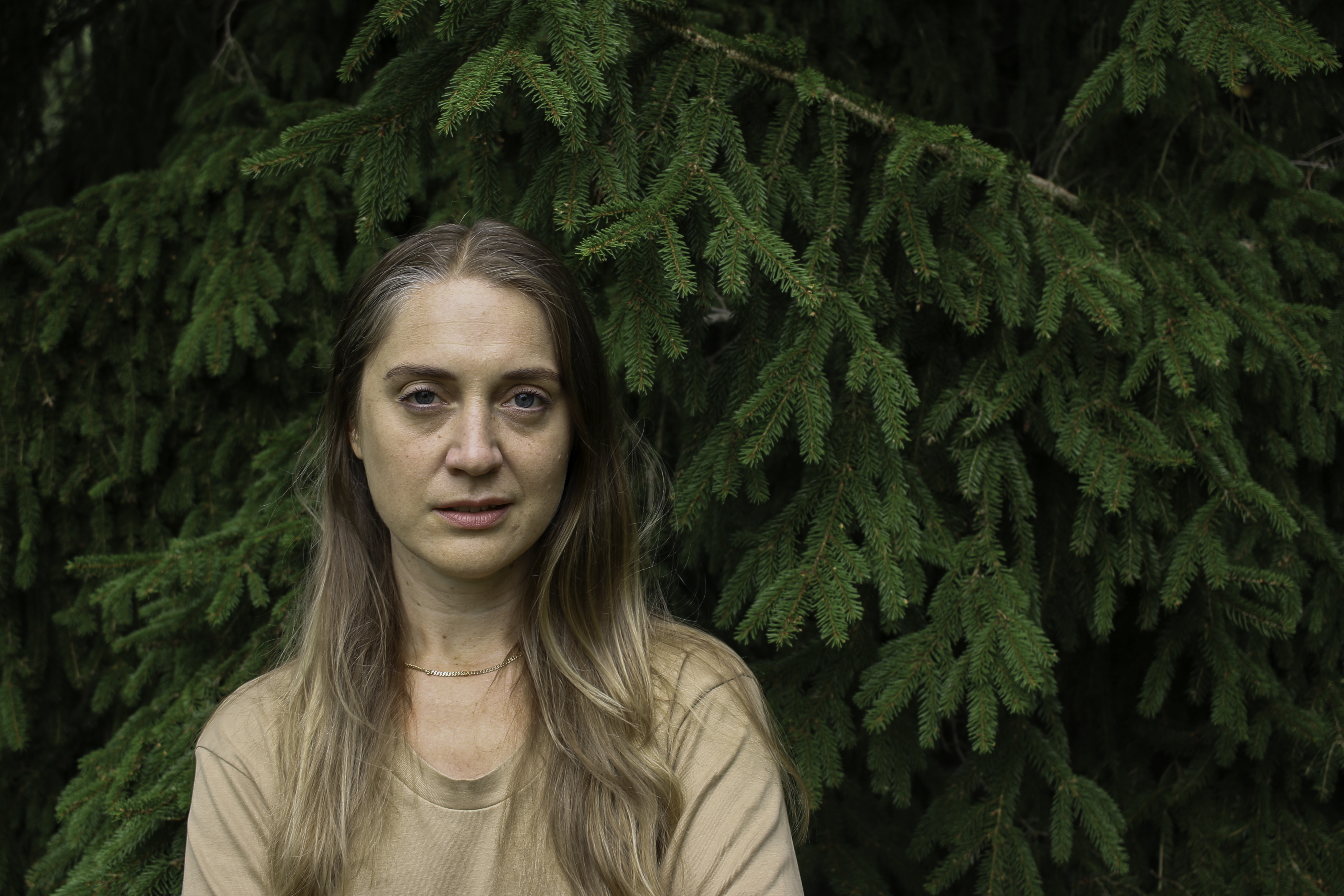 Konstnären Hanna Wildow står framför en grön barrskog. Hon har beige t-shirt och långt ljusbrunt hår.