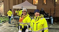 Anna och Jenny som arbetar på SMUAB står framför kameran och ler. De har på sig varseljackor och mössa och håller i de påsar som delades ut under inflyttningsfesten. I bakgrunden står ett vitt tält med SMUAB:s logga. Tältet är klätt med ljusslingor och i tältet står bor uppdukade med frukost.