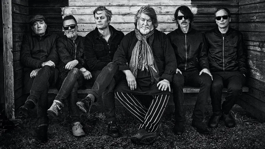 6 män sitter i mörka kläder på en bänk och tittar in i kameran. Bilden är svartvit.