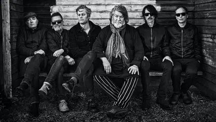 6 män sitter i mörka kläder på en bänk och tittar in i kameran. Bilden är svartvit.