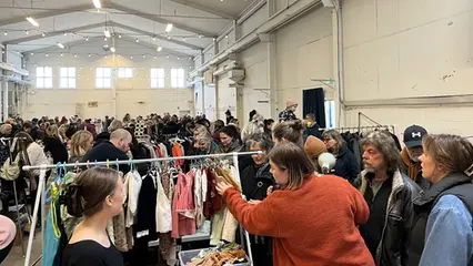 Människor i ett stort rum. En kvinna i röd jacka tittar bland kläderna på en klädställning.