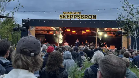 Ett publikhav framför en scen. Ovanpå scenen lyser en neonskylt med texten "Sommar i Skeppsbron".