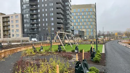 En gungställning står på en grön konstgräsmatta. I förgrunden syns planteringar av buskar och i bakgrunden syns ett svart hus med balkonger.