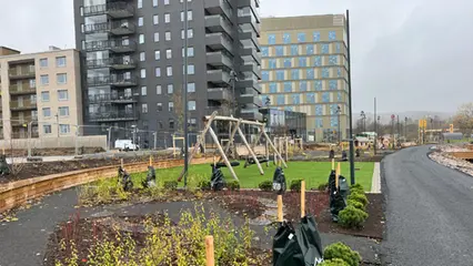 En gungställning står på en grön konstgräsmatta. I förgrunden syns planteringar av buskar och i bakgrunden syns ett svart hus med balkonger.