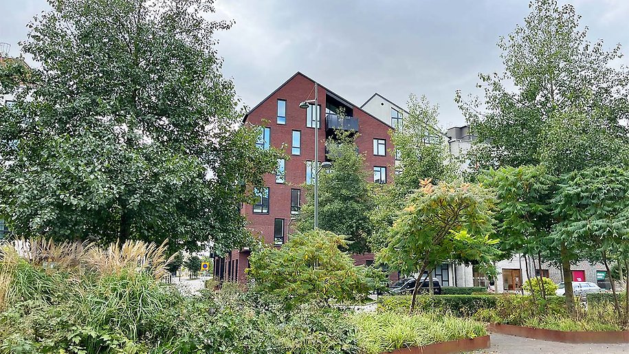 Ett rött tegelhus skymtar bakom gröna träd och planteringar i en park